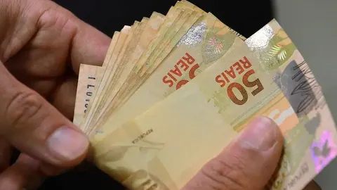 Mão segurando um maço de cédulas de 50 reais.